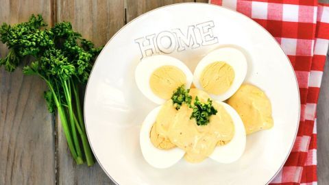 Auf einem weißen Teller mit dem Schriftzug "HOME" liegen halbe Eier, übergossen mit einer cremigen Senfsoße, garniert mit Frühlingszwiebeln. Neben dem Teller liegt ein kariertes полотенце.