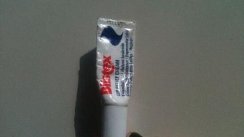 Blistex Lippencreme kann auch bei Pickeln helfen! Der Artikel beschreibt, wie eine kleine Tube dieser Creme über Nacht Hautunreinheiten reduzieren kann. Einfach auftragen und am nächsten Morgen saubere Haut genießen. Ein Blistex-Tuben wird gehalten, um Pickel zu behandeln, auf einer neutralen Oberfläche ohne weitere Elemente im Hintergrund.