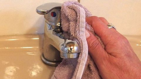 Eine Hand reinigt eine metallische Armatur mit einem Tuch, um verkalkte Armaturen zu vermeiden, im Badezimmer.