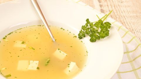 Ein klarer Eierstich in einer weißen Schüssel mit kleinen Würfeln und Kräutern wird mit einem Löffel serviert.