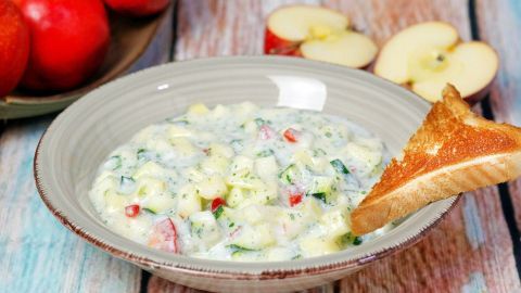Eine cremige Apfelsuppe wird in einer Schüssel serviert, während geröstetes Brot zum Dippen bereitliegt. Im Hintergrund sind frische Äpfel sichtbar.