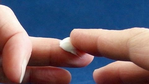 Zwei Finger berühren sich sanft, während ein kleines Stück weißer Substanz auf einem Finger sichtbar ist, vor blauem Hintergrund.