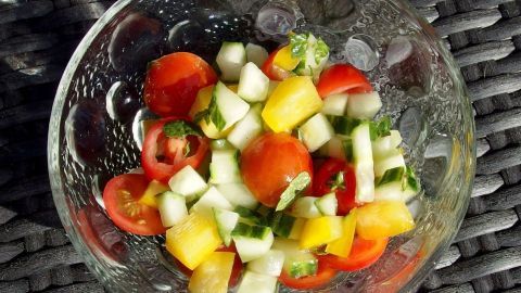 Frischer Salat in einer Glasschüssel, der aus Tomaten, Gurken und Paprika besteht, steht auf einem geflochtenen Untergrund.