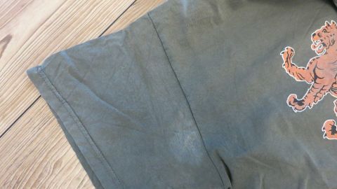 Ein grünes T-Shirt mit Deospuren auf dem Ärmel liegt auf einem Holzfußboden und zeigt ein farbiges Motiv.