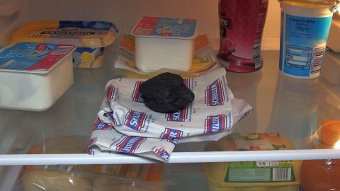 Eine schwarze, verschimmelte Lebensmittelverpackung liegt auf einem Handtuch im Kühlschrank, umgeben von verschiedenen Essensbehältern.