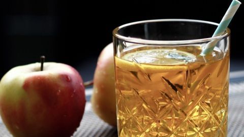 Ein Glas Apfelkornschorle steht auf einem Tisch, umgeben von frischen Äpfeln, während ein Strohhalm im Getränk steckt.