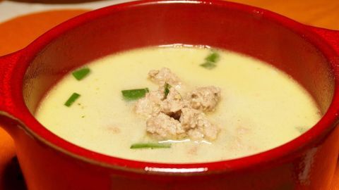 Eine rote Schüssel mit Käsesuppe steht auf einem bunten Tisch. Die Suppe ist cremig, mit Kräutern und kleinen Stückchen darüber.