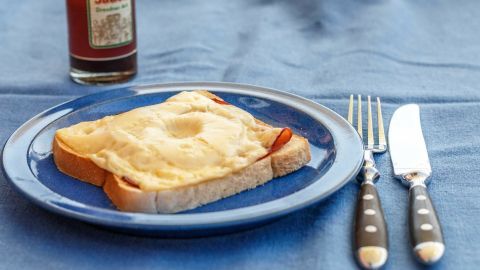 Ein belegtes Brot mit einer cremigen Schicht bedeckt liegt auf einem blauen Teller, umgeben von Besteck und einer Flasche Sauce.