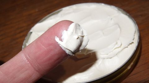 Eine Fingerkuppe ist mit Penaten-Creme bedeckt, die aus einem offenen Glasbehälter auf einem Tisch entnommen wurde.
