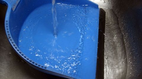 Fegen bei Wind: Vorher die Müllschaufel unter Wasser halten. Die feuchte Oberfläche bindet alles, was aufgefegt wird. Ein blauer Besenaufsatz steht in einer Spüle, während Wasser von oben hineinläuft, möglicherweise zum Reinigen von Balkon oder Terrasse.