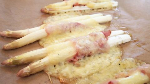 Weißer Spargel ist mit Schinken und geschmolzenem Käse überbacken, auf einem Backpapier in einer warmen Küche serviert.