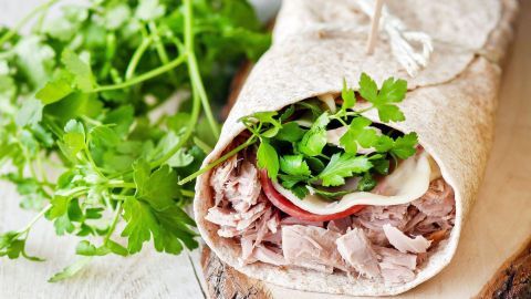 Ein nahrhafter Wrap aus Vollkorn mit Thunfisch, Käse und frischem Koriander liegt auf einem Holzbrett, umgeben von Kräutern.