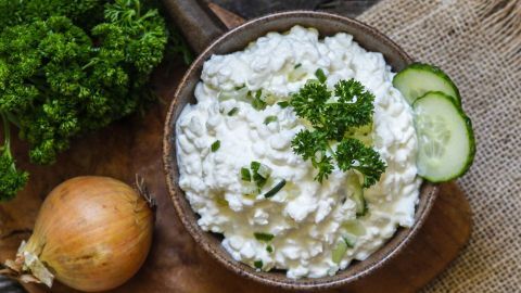 Eine Schüssel mit pikanter Frischkäse-Dip steht auf einem Holzbrett, umgeben von frischer Petersilie und einer Zwiebel.