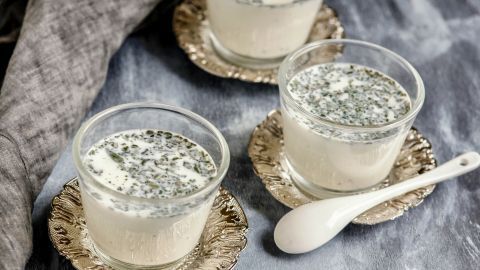 Drei Gläser Kräutermilch stehen auf glänzenden Untertellern, umgeben von einem grauen Hintergrund und einem Löffel.