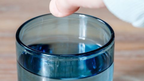 Eine Hand berührt die Oberfläche von blau gefärbtem Wasser in einem Glas, um das Orangeschimmer nach dem Haare färben zu demonstrieren.