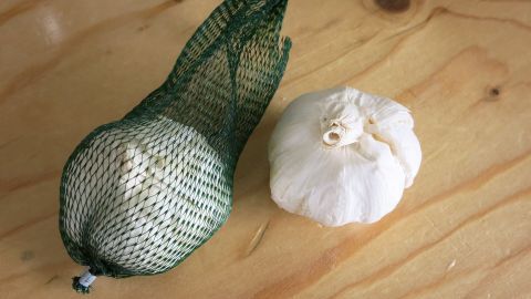 Leichtes Schälen von Knoblauch mithilfe eines Gummiverschlusses von einem Glasballon. Eine Knoblauchzehe liegt auf einem Holzuntergrund neben einer verpackten Knoblauchknolle, bereit zum Schälen.