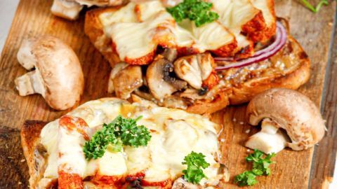 Überbackene Toastscheiben mit Putenschnitzel und Pilzen werden liebevoll garniert und serviert auf einem Holzbrett.