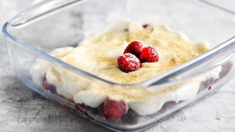 Ein klarer Glasbehälter zeigt einen French Kiss Nachtisch, üppig bedeckt mit Sahne und Himbeeren, auf einem stilvollen Tisch.