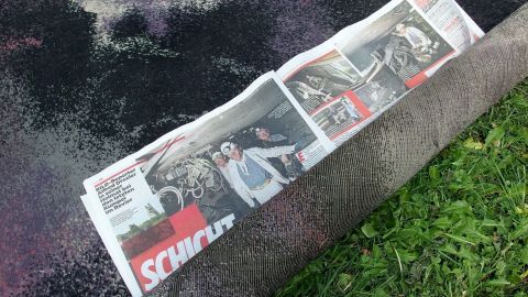 Mithilfe von Zeitungspapier Teppiche mottensicher lagern. Ein Teppich liegt auf dem Rasen, eingerollt mit Zeitungspapier zum Schutz vor Motten. Die Umgebung ist grün.