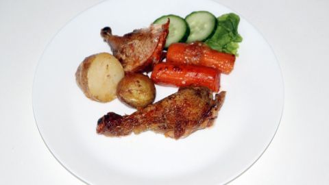 Saftiges Huhn liegt auf einem weißen Teller, umgeben von Kartoffeln, Karotten, Gurken und Blattsalat, appetitlich angerichtet.