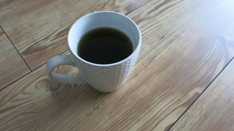 Eine Tasse schwarzer Tee steht auf einem glänzenden Parkettboden und reflektiert das Licht der Umgebung.