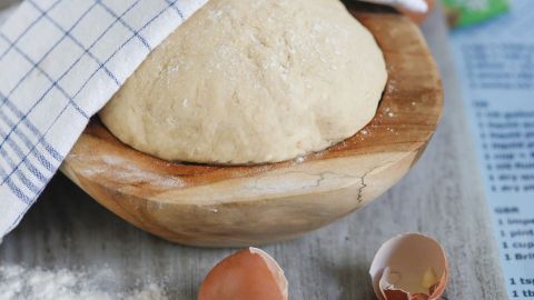 Ein Holzschüssel enthält einen ruhenden Hefeteig, bedeckt mit einem Küchenhandtuch, umgeben von Eierschalen und Mehl.