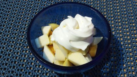 Eine Schüssel mit Obstsalat aus gewürfelten Äpfeln, bedeckt mit einem Häufchen Sahne, steht auf einem blauen Tisch.