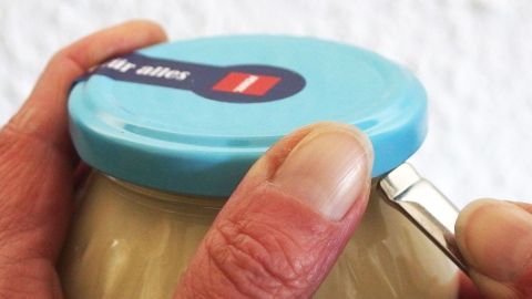Ein Glas wird ge&ouml;ffnet, w&auml;hrend eine Hand mit einem Werkzeug den Deckel anhebt. Der Hintergrund ist hell und neutral.