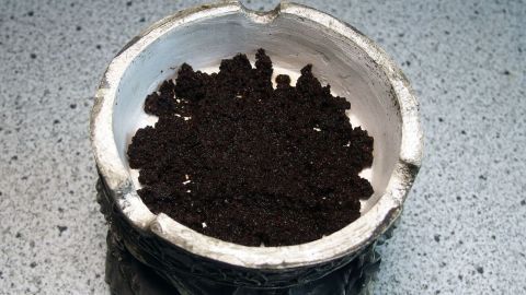 In den Boden des Aschenbechers eine dünne Schicht gemahlenen Kaffee geben. Ein Aschenbecher aus natürlichem Material enthält Kaffeemehl und steht auf einer glatten Oberfläche in einer modernen Umgebung.