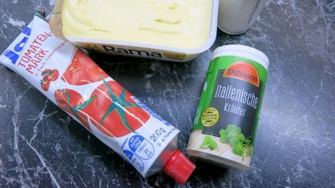 Tomatenbutter wird zubereitet, indem eine Butterdose, Tomatenpaste und Gewürze auf einer dunklen Arbeitsfläche angeordnet sind.