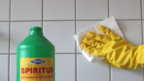 Eine grüne Flasche mit Spiritus steht neben einer Hand in einem gelben Gummihandschuh, die eine Fläche abwischt.