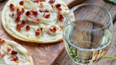 Zwei Elsässer Flammkuchen mit Speck liegen auf einem Holzbrett, während ein elegantes Weinglas daneben steht.