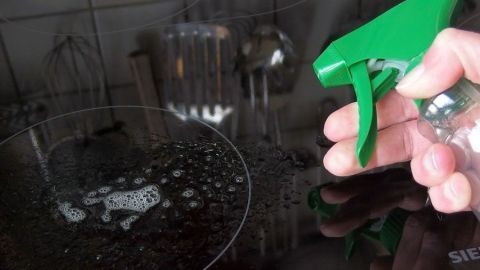 Ein Hand sprüht Glasreiniger auf ein Glaskeramikfeld, um es strahlend sauber zu machen. Im Hintergrund sind Küchenutensilien sichtbar.