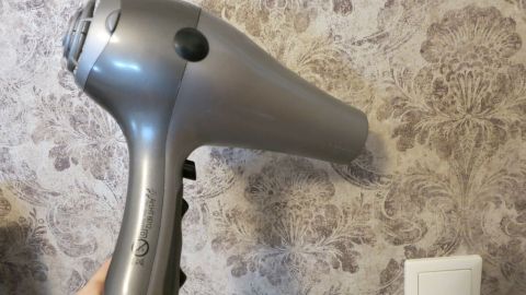 So entfernt man Wachsflecken auf der Tapete: Mit dem Föhn das Wachs flüssig machen und dabei laufend mit dem Microfasertuch aufwischen, bis keine Wachsspuren bleiben! Ein Föhn wird aktiv auf eine Tapete gerichtet, um Wachsflecken zu entfernen. Der Hintergrund zeigt ein blumiges Tapetenmuster.