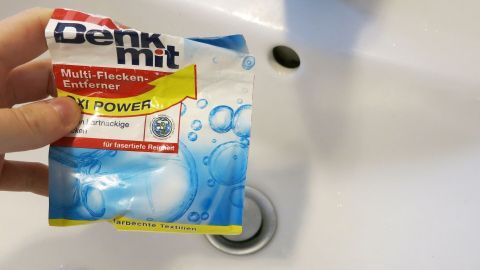 Ein Hand hält ein Paket Fleckensalz über einem weißen Spülbecken, bereit zur Reinigung. Der Wasserhahn ist sichtbar.