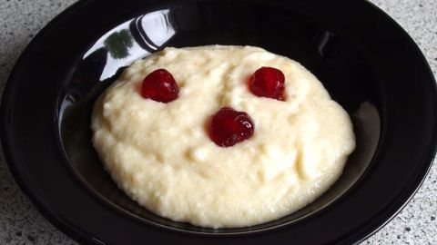 Ein schwarzes Schälchen mit Grießbrei, in dem rote Kirschen platziert sind, steht auf einer hellen Arbeitsplatte.