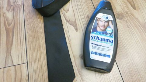 Fettflecken auf Krawatten lassen sich einfach mit Schuppenshampoo behandeln. Eine schwarze Krawatte liegt neben einem Fläschchen Schuppenshampoo auf einem Holzfußboden, bereit zur Fleckenentfernung.