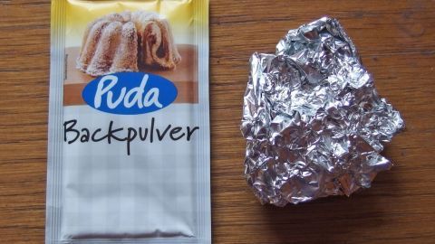 Ein Päckchen Backpulver liegt neben einer zerknüllten Alufolie auf einer Holzoberfläche, bereit für die Reinigung von Herdplatten.