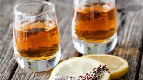 Zwei Gläser Nikolaschka stehen auf einem rustikalen Holztisch, umgeben von Zitronenscheiben mit Salz und Gewürzen.