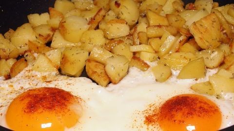 In einer Pfanne braten zwei Spiegeleier zusammen mit knusprigen Kartoffelstücken, die mit Gewürzen verfeinert sind.
