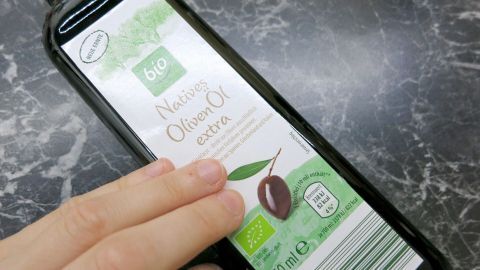 Olivenöl kann spröde Nägel stärken! Massiere regelmäßig erwärmtes Öl in deine Nägel und Nagelhaut ein, um sie zu pflegen und Spliss zu reduzieren. Die Aufnahme zeigt eine Hand, die eine Flasche Bio-Olivenöl hält. Eine Hand berührt eine Flasche mit dem Etikett "Bio Natives Olivenöl extra" auf einer dunklen Marmoroberfläche.