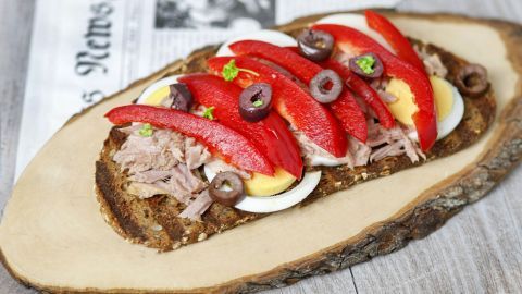 Ein belegtes Brot mit Thunfisch, Eiern, Paprika und Oliven liegt auf einem Holzbrett, umgeben von einem neutralen Hintergrund.