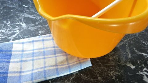 Damit du die Masse in der Schüssel ohne Verrutschen selbiger umrühren kannst, kannst du darunter einen feuchten Lappen legen. Ein gelber Rührschüssel steht auf einer dunklen, marmorierten Oberfläche, während ein Tuch darunter liegt, das ein Verrutschen verhindert.