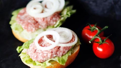 Auf zwei Brötchen liegt Hackepeter, garniert mit Zwiebeln und frischem Salat, neben zwei glänzenden Tomaten.
