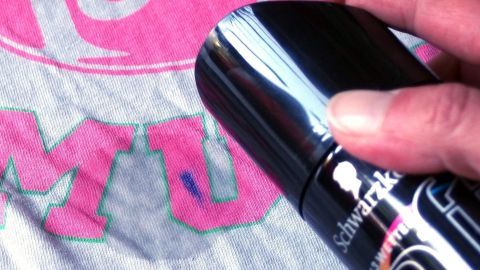 Flecken von Filzstift auf Kleidung mithilfe von Haarspray entfernen. Ein Hand hält einen Fleckenentferner über ein T-Shirt mit Filzstiftflecken, das auf einem hellen Hintergrund liegt.