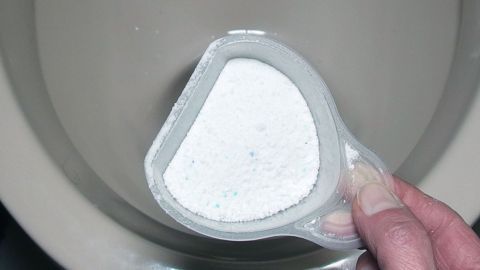 Eine Hand hält ein transparentes Gefäß mit Waschpulver über einer Toilettenschüssel, bereit zum Einsatz.
