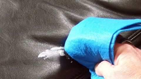Ein Handtuch reinigt einen Lederpolster, während Wasser darauf aufgetragen wird, um die Oberfläche zu pflegen und aufzufrischen.