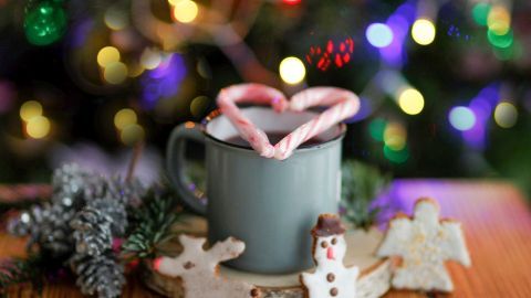 Eine graue Tasse mit Pfefferminzstäbchen in Herzform steht auf einem Holzbrett, umgeben von Keksen und festlichem Hintergrund.