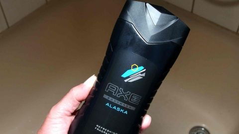 Ein schwarzer Shampoo-Flakon wird in einer Hand gehalten, während im Hintergrund eine Dusche oder Badewanne zu sehen ist.