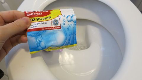 Mithilfe von Fleckensalz lässt sich das Toilettenbecken hervorragend reinigen. Ein Hand hält ein Päckchen Fleckensalz über ein sauberes Toilettenbecken, bereit zur Anwendung zur Reinigung.